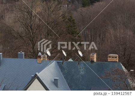 Residential grey roofs feature multiple satellite dishes and brick chimneys, symbolizing modern communication and home connectivity in a winter forest Residential grey roofs feature multiple satellite dishes and brick chimneys, symbolizing modern communication and home connectivity in a winter forest 139412567