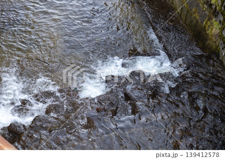 Dynamic stream water vigorously flowing over dark river rocks, generating frothy white bubbles, representing nature's refreshing purity and ecological vitality for environmental concepts Dynamic stream water vigorously flowing over dark river rocks, generating frothy white bubbles, representing nature's refreshing purity and ecological vitality for environmental concepts 139412578