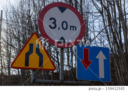 Road signs on a pole display a 3,0 m height limit, road narrowing, and traffic priority, guiding driver safety and efficient urban navigation 139412585