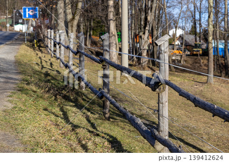 Rustic wooden fence with rough logs and wire defines a sunny country road boundary, leading towards distant village houses and bare forest trees Rustic wooden fence with rough logs and wire defines a sunny country road boundary, leading towards distant village houses and bare forest trees 139412624