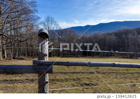 Rustic wooden fence with electric wire marking a winter field boundary, showcasing bare trees and distant mountains under a bright blue sky, portraying rural nature Rustic wooden fence with electric wire marking a winter field boundary, showcasing bare trees and distant mountains under a bright blue sky, portraying rural nature 139412625
