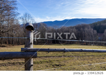 Weathered wooden fence with electric wires delineating a calm rural field, featuring bare winter trees and distant mountains under a blue sky Weathered wooden fence with electric wires delineating a calm rural field, featuring bare winter trees and distant mountains under a blue sky 139412626