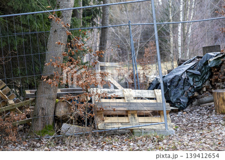 Metal construction fence leaning against a tree with wooden pallets and dry leaves on the ground, illustrating an abandoned building site or outdoor development Metal construction fence leaning against a tree with wooden pallets and dry leaves on the ground, illustrating an abandoned building site or outdoor development 139412654