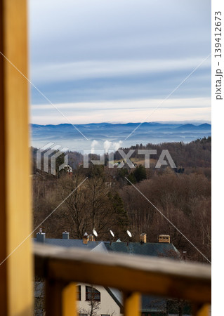 Panoramic mountain landscape vista with misty valleys, bare winter trees, distant industrial smoke, and rooftops, framed by a wooden balcony railing Panoramic mountain landscape vista with misty valleys, bare winter trees, distant industrial smoke, and rooftops, framed by a wooden balcony railing 139412673