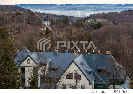 Traditional European resort buildings with grey roofs, satellite dishes, amidst bare winter trees, overlooking a serene, hazy blue mountain range Traditional European resort buildings with grey roofs, satellite dishes, amidst bare winter trees, overlooking a serene, hazy blue mountain range 139412683