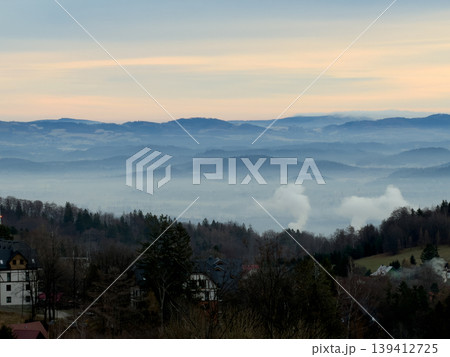Panoramic mountain range view with a hazy valley, scattered houses, and industrial smoke plumes, depicting both serene nature and human influence Panoramic mountain range view with a hazy valley, scattered houses, and industrial smoke plumes, depicting both serene nature and human influence 139412725