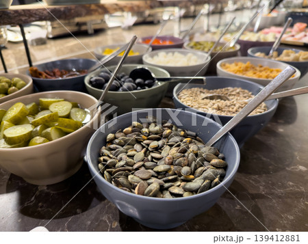 Buffet bar featuring various bowls of healthy seeds, olives, and pickles, offering diverse nutritious options for a hotel breakfast or salad Buffet bar featuring various bowls of healthy seeds, olives, and pickles, offering diverse nutritious options for a hotel breakfast or salad 139412881