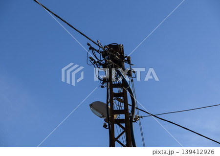 Rusted electricity pole featuring a complex web of tangled power lines and an urban street light, conveying essential energy infrastructure against a clear blue sky 139412926