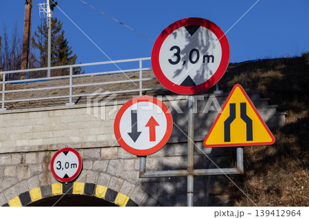 Multiple road signs display a 3,0 m height restriction under a bridge, alongside warnings for narrow passages and traffic priority, promoting road safety 139412964