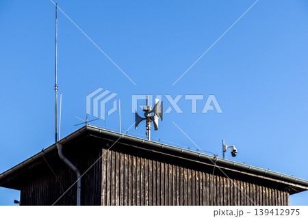 Rustic wooden building roof featuring a public safety siren, various communication antennas, and a security camera, essential for warning and surveillance Rustic wooden building roof featuring a public safety siren, various communication antennas, and a security camera, essential for warning and surveillance 139412975