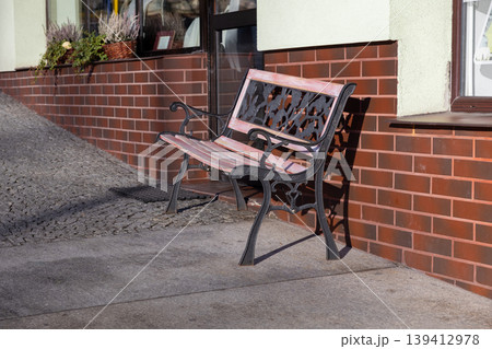 Vintage wrought iron bench with light pink wooden slats casts a sharp shadow on the sidewalk by a textured brick wall, offering public relaxation and urban charm Vintage wrought iron bench with light pink wooden slats casts a sharp shadow on the sidewalk by a textured brick wall, offering public relaxation and urban charm 139412978