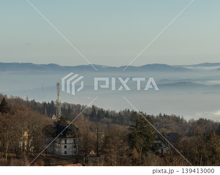 Mountain landscape with a communication tower rising above houses and a valley filled with dense fog, creating a serene and expansive morning view Mountain landscape with a communication tower rising above houses and a valley filled with dense fog, creating a serene and expansive morning view 139413000