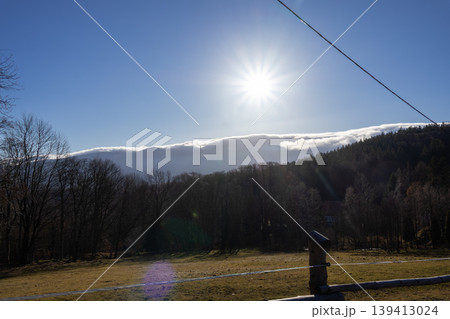 Bright sun with starburst effect shining over mountain peaks layered with white clouds and a lush forest, creating a stunning scenic natural landscape Bright sun with starburst effect shining over mountain peaks layered with white clouds and a lush forest, creating a stunning scenic natural landscape 139413024