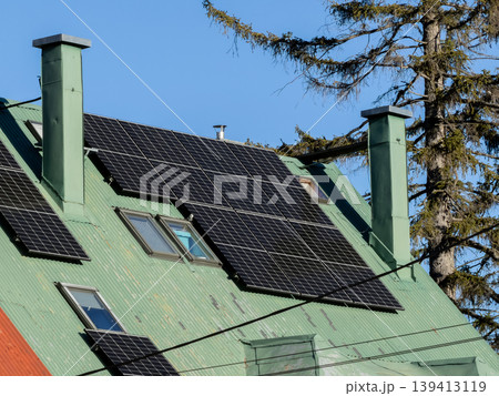 A solar panel array is mounted on a green corrugated metal roof with chimneys, representing residential renewable energy, sustainable living, clean power infrastructure, and eco-friendly home upgrades 139413119
