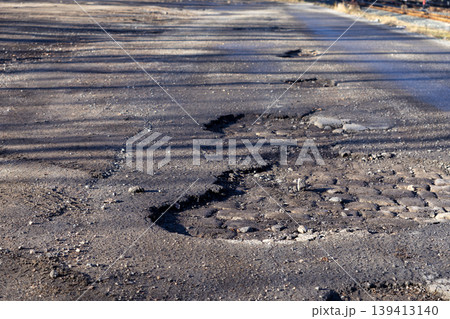 Damaged asphalt road with deep potholes and cracked surface, illustrating infrastructure decay, lack of maintenance, and unsafe driving conditions in an urban area or along a neglected rural highway Damaged asphalt road with deep potholes and cracked surface, illustrating infrastructure decay, lack of maintenance, and unsafe driving conditions in an urban area or along a neglected rural highway 139413140