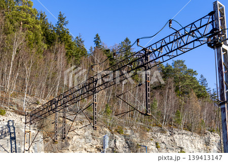 A modern catenary system spans across a railway track nestled against a rugged rocky hillside and lush forest, illustrating efficient transportation infrastructure and precise engineering for travel A modern catenary system spans across a railway track nestled against a rugged rocky hillside and lush forest, illustrating efficient transportation infrastructure and precise engineering for travel 139413147