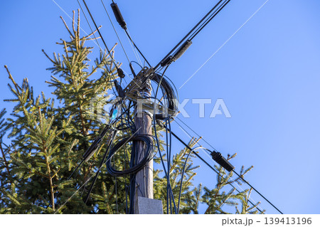 A weathered wooden utility pole supporting a complex tangle of electrical power lines and communication cables stands against a bright clear blue sky, illustrating vital urban and rural connectivity A weathered wooden utility pole supporting a complex tangle of electrical power lines and communication cables stands against a bright clear blue sky, illustrating vital urban and rural connectivity 139413196