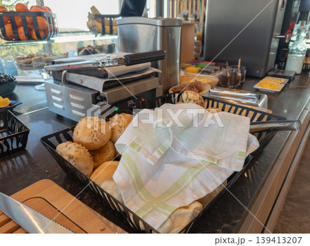 Fresh rustic bread rolls in a basket covered with a kitchen cloth, arranged on a hotel buffet table to offer a gourmet breakfast experience, promoting hospitality, travel, and culinary convenience 139413207