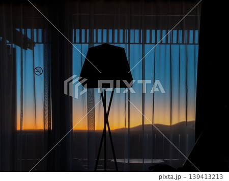 Floor lamp silhouette against window with curtains, capturing a vibrant golden hour sunset over distant mountains to represent calm, relaxation, and luxury interior design concepts in a modern hotel Floor lamp silhouette against window with curtains, capturing a vibrant golden hour sunset over distant mountains to represent calm, relaxation, and luxury interior design concepts in a modern hotel 139413213