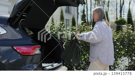Senior woman puts bag in car trunk and closes it with button. Senior woman puts bag in car trunk and closes it with button. 139413778