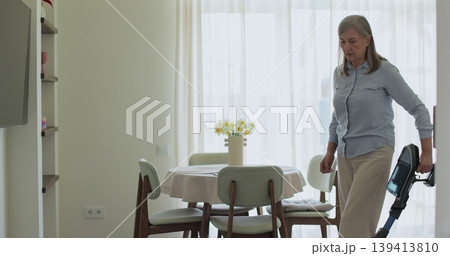 Senior woman cleans apartment by hand vacuuming carpet in living room. Concept of daily home routine. 139413810
