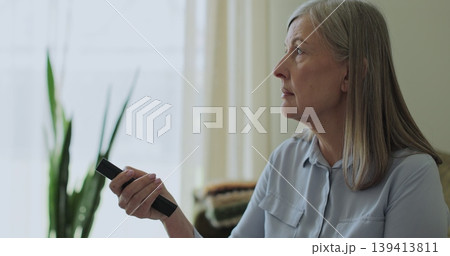 Elder woman selects tv channels with remote control at living room on sofa. Woman controls TV using a modern remote control. Elder woman selects tv channels with remote control at living room on sofa. Woman controls TV using a modern remote control. 139413811