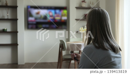 Elder woman selects tv channels with remote control at living room on sofa. Woman controls TV using a modern remote control. Elder woman selects tv channels with remote control at living room on sofa. Woman controls TV using a modern remote control. 139413819