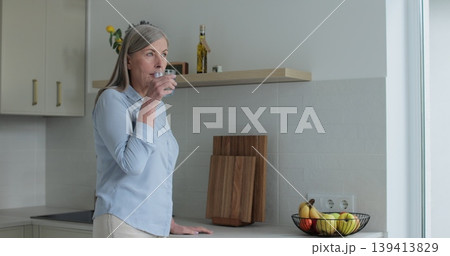 Senior mature woman drinking a glass of water in kitchen at home. Elderly, lifestyle, hydration, wellness, relaxation, home. Senior mature woman drinking a glass of water in kitchen at home. Elderly, lifestyle, hydration, wellness, relaxation, home. 139413829