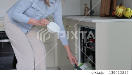 Senior woman unloading dishwasher at home. Mature housewife doing household taking clear dishes from dishwasher in modern kitchen. Senior woman unloading dishwasher at home. Mature housewife doing household taking clear dishes from dishwasher in modern kitchen. 139413835