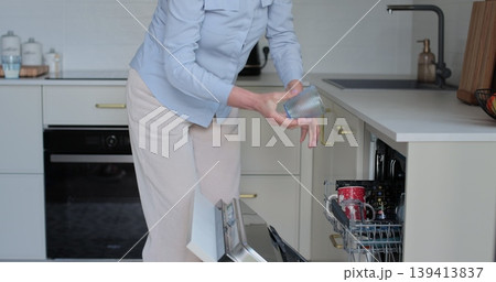 Senior woman unloads clean dishes from dishwasher machine. Concept of using kitchen appliances. Senior woman unloads clean dishes from dishwasher machine. Concept of using kitchen appliances. 139413837