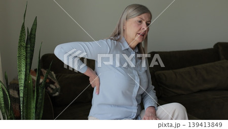 Mature woman feeling ache lower back pain on sofa at home. Upset elderly retired woman massages painful lower back muscles. Senior lady feel spasm. 139413849