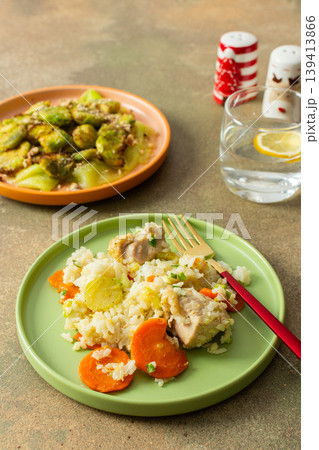 Boiled rice topped with chicken breast serves as the main dish. A side of Brussels sprouts and zucchini slices is presented. The setting includes a flower and seasonings Boiled rice topped with chicken breast serves as the main dish. A side of Brussels sprouts and zucchini slices is presented. The setting includes a flower and seasonings 139413866