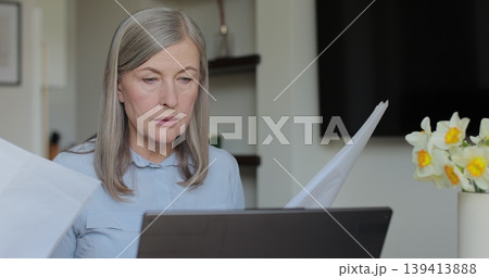 Senior woman sits at table viewing papers and utility bills. Senior woman sit with laptop and paper document. Senior woman sits at table viewing papers and utility bills. Senior woman sit with laptop and paper document. 139413888