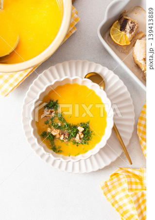 Pumpkin cream soup sits in a bowl with toppings next to a dish of baked hake and lemon. A spoon rests beside the soup in a clean dining area 139413889
