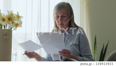 Close up older gray-haired woman sits at a table in the living room, calculating bills and taxes and taking notes. Close up older gray-haired woman sits at a table in the living room, calculating bills and taxes and taking notes. 139413933