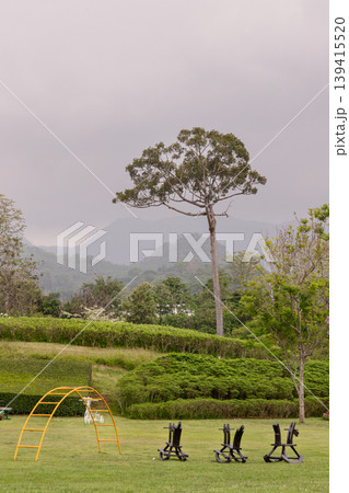 Mist clings to the rolling mountains as a sprawling emerald lawn unfolds under a heavy sky. Here, dark sculptures and a solitary golden climbing frame stand in quiet contrast to the lush surroundings. Mist clings to the rolling mountains as a sprawling emerald lawn unfolds under a heavy sky. Here, dark sculptures and a solitary golden climbing frame stand in quiet contrast to the lush surroundings. 139415520