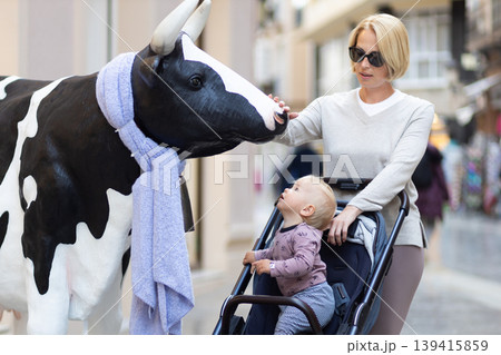 CuriouMother and little son exploring city streets and discovering a funny cow sculptures toddler in a stroller looking at a cow statue in front of a shop in Spain. 139415859