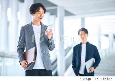 スマートフォンを見る30代男性と後方の50代男性のビジネスシーン 139416187
