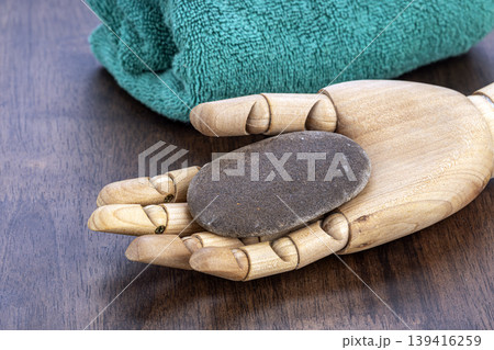 Wooden Articulated Mannequin Hand Holding Wet Smooth Pumice Stone to Exfoliate Dry Skin with Green Towel on Dark Wood Surface Wooden Articulated Mannequin Hand Holding Wet Smooth Pumice Stone to Exfoliate Dry Skin with Green Towel on Dark Wood Surface 139416259
