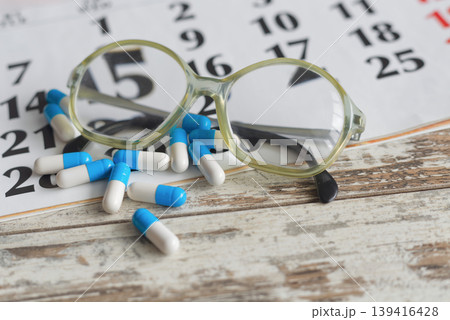 White and blue pills and reading glasses lie on a large paper calendar on an old wooden table, for treatment strategy, path to health, and medical control themes. White and blue pills and reading glasses lie on a large paper calendar on an old wooden table, for treatment strategy, path to health, and medical control themes. 139416428