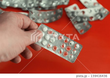 Close-up of a hand holding the last half-empty blister pack of pills against a pile of used empty packages on a bright red background. Close-up of a hand holding the last half-empty blister pack of pills against a pile of used empty packages on a bright red background. 139416436