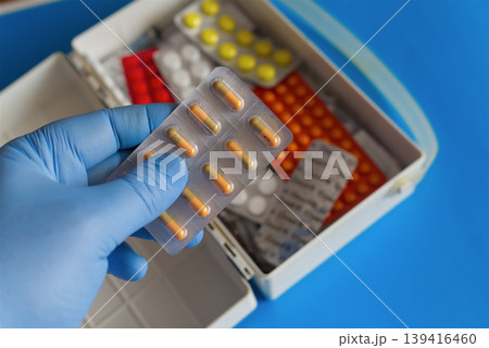 Hand in a blue medical glove holding a blister with colorful pills against the background of an open first aid kit with various colorful tablets, close-up. Hand in a blue medical glove holding a blister with colorful pills against the background of an open first aid kit with various colorful tablets, close-up. 139416460
