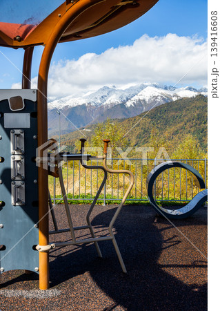 Outdoor fitness equipment located on a mountain. The area offers a view of snow-capped peaks and green hills under a blue sky with clouds. Outdoor fitness equipment located on a mountain. The area offers a view of snow-capped peaks and green hills under a blue sky with clouds. 139416608