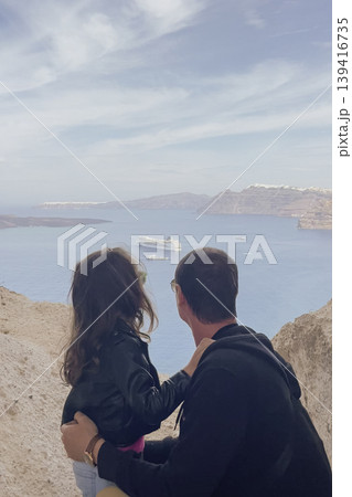 Father and daughter sitting on Santorini cliff edge, embracing and looking at Aegean Sea, caldera, anchored cruise ship, surrounding white cliffside houses Father and daughter sitting on Santorini cliff edge, embracing and looking at Aegean Sea, caldera, anchored cruise ship, surrounding white cliffside houses 139416735