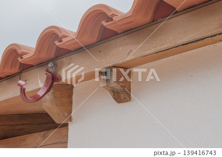 A detailed shot of water damage on a wooden roof beam due to a missing gutter system. The image shows wood rot, darkening, and a lone metal bracket on a tiled roof A detailed shot of water damage on a wooden roof beam due to a missing gutter system. The image shows wood rot, darkening, and a lone metal bracket on a tiled roof 139416743