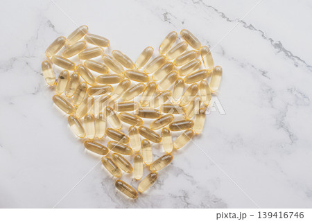 A professional overhead shot of golden Omega-3 fish oil capsules arranged in a heart shape on a white marble surface. The translucent pills glow in soft light 139416746