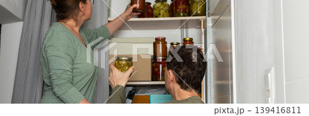 Married couple places jars of canned goods onto shelves in their pantry. They are inside their home with a view of flowers and the city from the balcony. It is autumn, banner 139416811