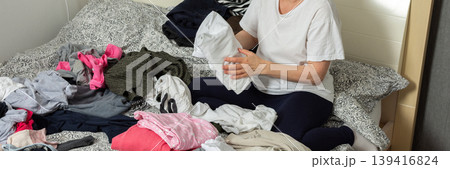 A 52-year-old woman is sorting clean laundry on a bed. She holds a pair of pants in one hand while looking at the clothes around her. A plant is seen in the corner of the room, banner A 52-year-old woman is sorting clean laundry on a bed. She holds a pair of pants in one hand while looking at the clothes around her. A plant is seen in the corner of the room, banner 139416824