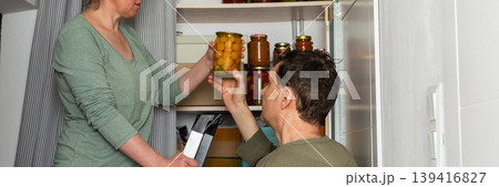 Married couple places jars of canned goods onto shelves in their pantry. They are inside their home with a view of flowers and the city from the balcony. It is autumn, banner Married couple places jars of canned goods onto shelves in their pantry. They are inside their home with a view of flowers and the city from the balcony. It is autumn, banner 139416827
