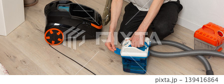 A 53-year-old man kneels on the floor as he prepares his vacuum cleaner for cleaning. He checks parts and organizes equipment in a well-lit room with plants, banner A 53-year-old man kneels on the floor as he prepares his vacuum cleaner for cleaning. He checks parts and organizes equipment in a well-lit room with plants, banner 139416864